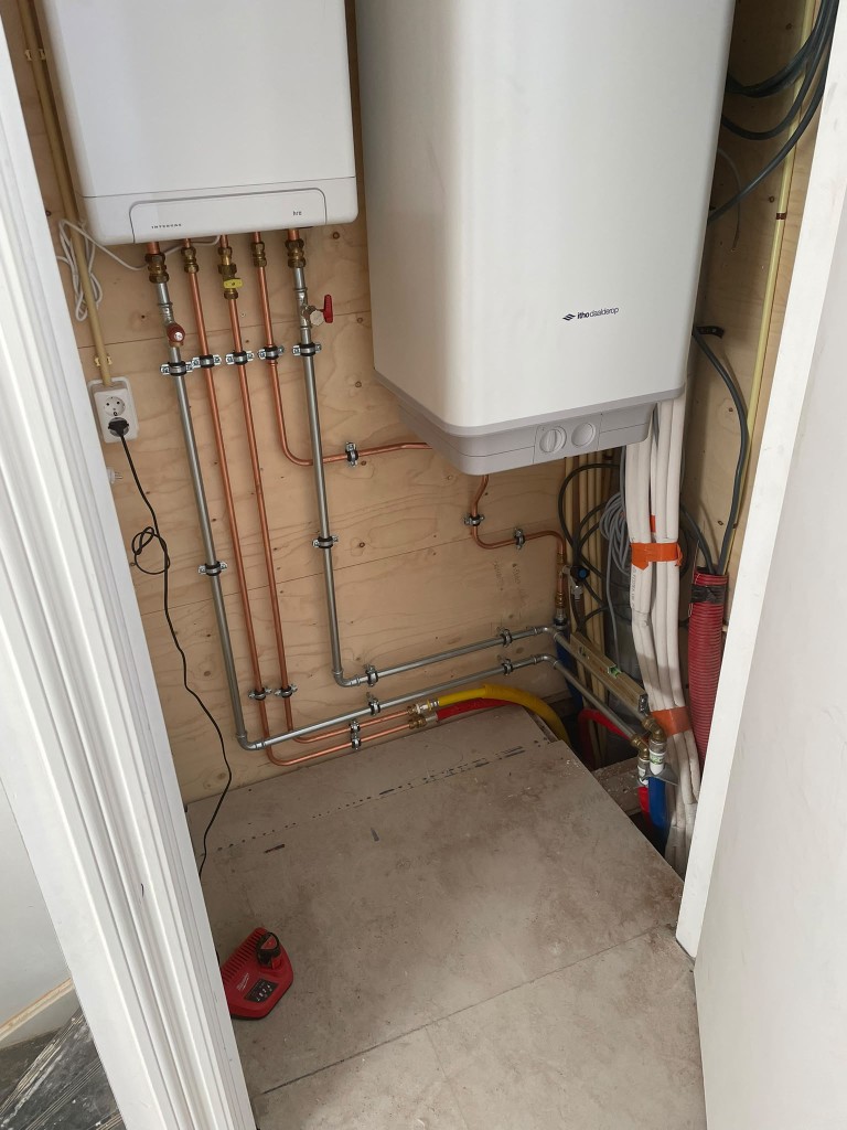 Koperen waterleidingen, afsluiters en twee installaties tegen een houten wand.